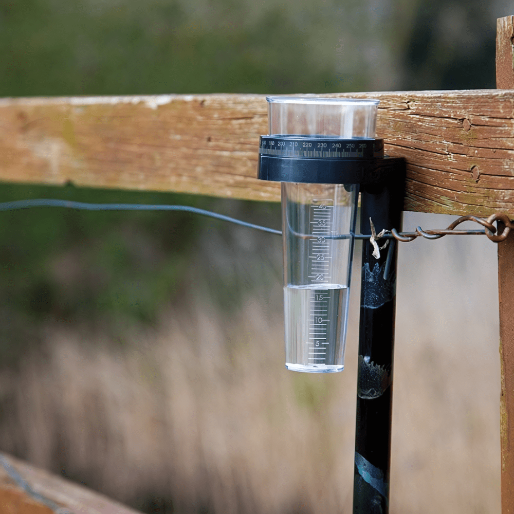 Un pluviomètre Thermomètre.fr fixé sur un poteau de clôture en bois mesurant les précipitations en milieu naturel.
